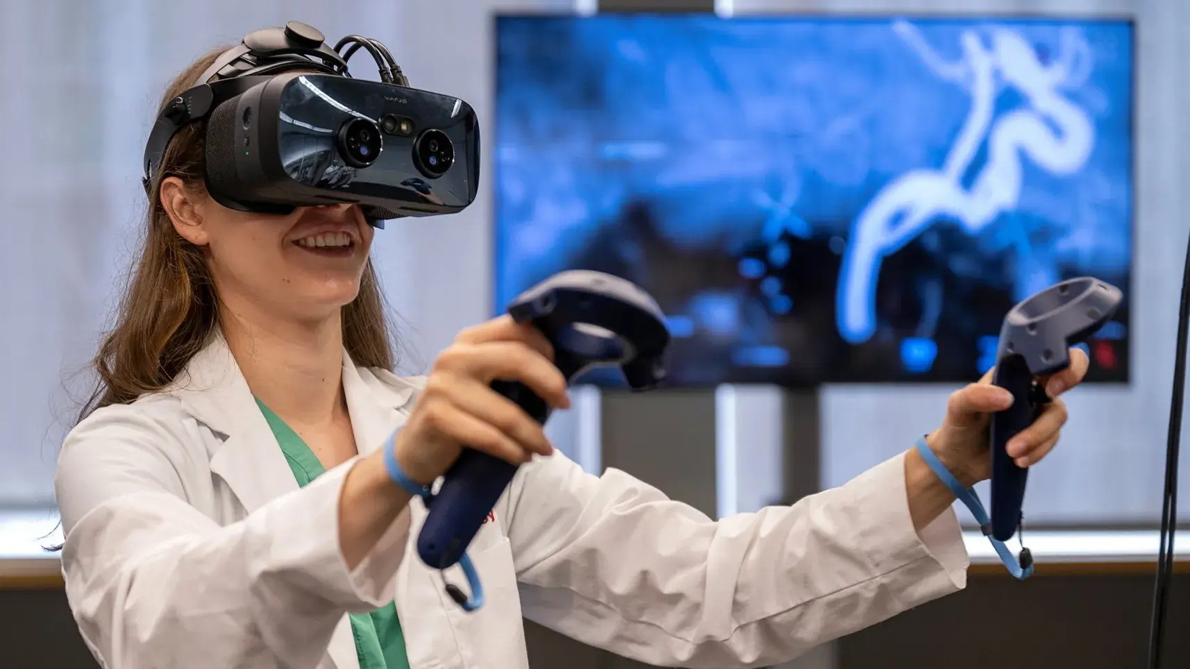 Female student in a lab coat, wearing VR headset and holding controllers, engaging with anatomy that is on a TV behind her.